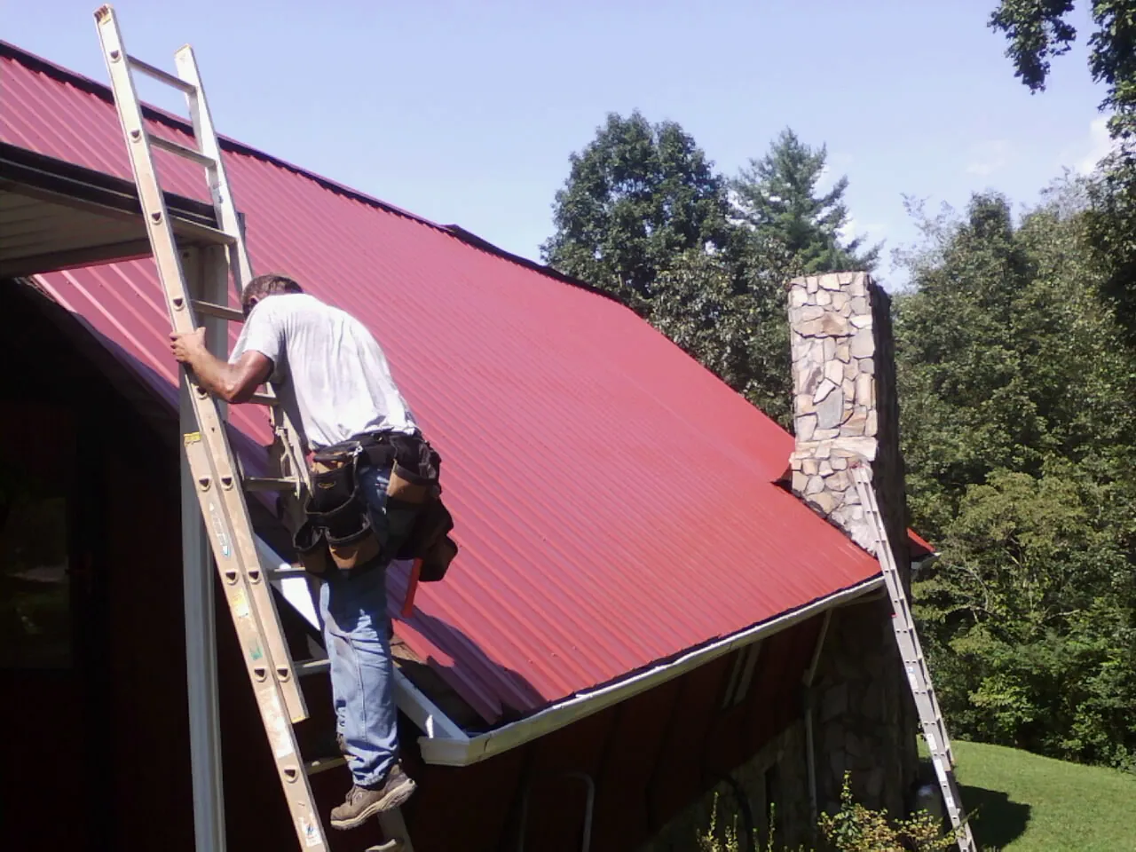 Roofer on a ladder beside a red metal roof and stone chimney — Soffit & Fascia Repair by Roofing Brothers in Savage